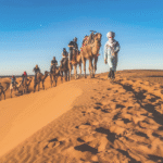 Camel tour in the Merzouga desert, Morocco, with travelers riding through golden sand dunes at sunset.